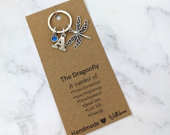 May include: A silver-toned metal keychain featuring a dragonfly charm, a letter A charm, and a small blue circular crystal charm attached to a ring. The keychain is mounted on a rectangular brown card with the printed text The Dragonfly A symbol of Transformation New beginnings Development Good luck Live life Change Handmade With love.