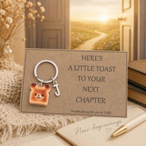 May include: A silver-toned metal keychain featuring a small, light brown bear-shaped toast charm and a letter J charm. The keychain is attached to a rectangular brown card with the text: HERE'S A LITTLE TOAST TO YOUR NEXT CHAPTER. Handmade just for you by TzUD.