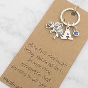 Puede incluir: Un llavero de plata con un dije de elefante, un dije de cristal azul y un dije de plata con la letra A. El llavero está unido a una tarjeta marrón con el texto "May this elephant bring you good luck, prosperity, strength and success in all you" y "Handmade".