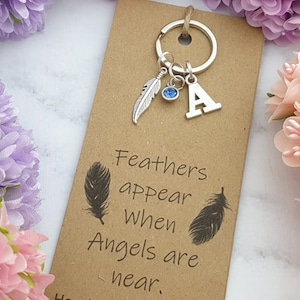 Puede incluir: Un llavero plateado con un dije de pluma, una gema azul y la letra "A" en una tarjeta marrón. La tarjeta dice "Las plumas aparecen cuando los ángeles están cerca". Flores decorativas rodean la tarjeta.