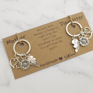 May include: Two silver keychains with charms, one with the word "Mother" and the other with the word "Daughter". Both keychains have a sunflower charm and a broken heart charm. The keychains are on a brown card with the text "We have been Mother & Daughter right from the start. The friendship we share is a gift from the heart. Handmade with love."