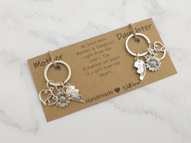 May include: Two silver keychains with charms, one with the word "Mother" and the other with the word "Daughter". Both keychains have a sunflower charm and a broken heart charm. The keychains are on a brown card with the text "We have been Mother & Daughter right from the start. The friendship we share is a gift from the heart. Handmade with love."