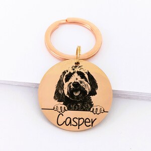 May include: A round, copper-coloured keychain with a black and white photo of a dog engraved on it. The dog is looking at the camera and has its paws on the edge of the keychain. The name "Casper" is engraved below the photo.