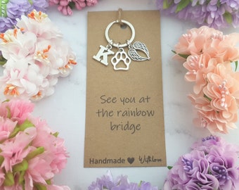 Puede incluir: Un llavero de metal plateado con un dije de la letra K, un dije de huella de pata y un dije de alas de ángel en una anilla. El llavero está montado en una tarjeta rectangular marrón con el texto See you at the rainbow bridge.