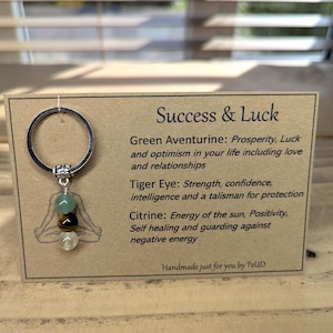 Könnte beinhalten: Ein Schlüsselanhänger mit einem silbernen Ring und einem Anhänger mit grünen, braunen und klaren Edelsteinen. Die dazugehörige Karte trägt die Aufschrift "Success & Luck" und listet die Eigenschaften der Edelsteine auf. Der Hintergrund ist eine Holzoberfläche.