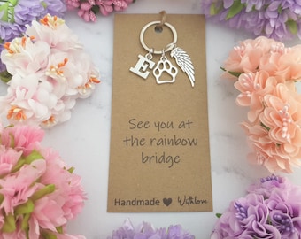 Puede incluir: Un llavero de metal plateado con un dije de letra E, un dije de huella de pata y un dije de ala de ángel en una argolla. El llavero está montado en una tarjeta marrón rectangular con el texto See you at the rainbow bridge y Handmade with love.