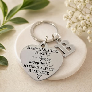 May include: A silver-toned metal keyring featuring a heart-shaped pendant and a separate letter B charm. The heart pendant is engraved with the black text: SOMETIMES YOU FORGET You're awesome SO THIS IS A LITTLE REMINDER, accented with two small heart icons.