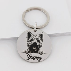May include: Silver key chain with a round tag featuring a black and white illustration of a Westie dog peeking over the top. The tag is engraved with the name "Darcy".
