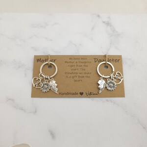 May include: Two brown cardstock tags with the words "Mother" and "Daughter" printed on them. Each tag has a silver key chain with charms including a sunflower, a heart, and a broken heart with the words "Best Friends" on it. The text on the tags reads "We have been Mother & Daughter right from the start. The friendship we share is a gift from the heart. Handmade Withlove."