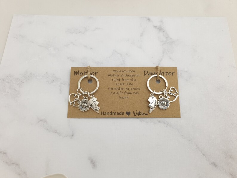 May include: Two brown cardstock tags with the words "Mother" and "Daughter" printed on them. Each tag has a silver key chain with charms including a sunflower, a heart, and a broken heart with the words "Best Friends" on it. The text on the tags reads "We have been Mother & Daughter right from the start. The friendship we share is a gift from the heart. Handmade Withlove."