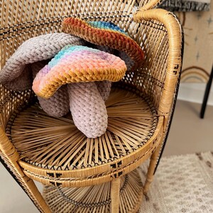 Pode incluir: Dois brinquedos de cogumelo arco-&iacute;ris de croch&ecirc; em uma cadeira de vime. Os cogumelos s&atilde;o feitos com um fio macio e felpudo e t&ecirc;m um esquema de cores de arco-&iacute;ris pastel.