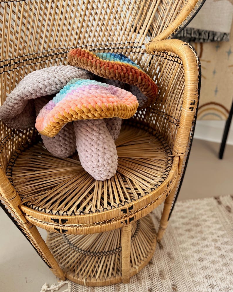 Pode incluir: Dois brinquedos de cogumelo arco-&iacute;ris de croch&ecirc; em uma cadeira de vime. Os cogumelos s&atilde;o feitos com um fio macio e felpudo e t&ecirc;m um esquema de cores de arco-&iacute;ris pastel.