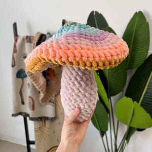 Pode incluir: Um brinquedo de cogumelo de croch&ecirc; com uma parte superior arco-&iacute;ris e um caule rosa.