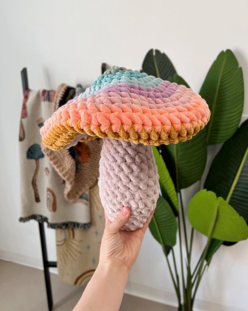 Pode incluir: Um brinquedo de cogumelo de croch&ecirc; com uma parte superior arco-&iacute;ris e um caule rosa.