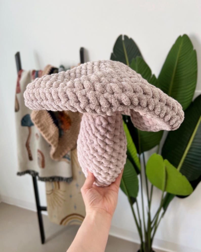Pode incluir: Um brinquedo de cogumelo de croch&ecirc; rosa macio e felpudo. O cogumelo tem uma tampa grande e redonda e um caule redondo menor.