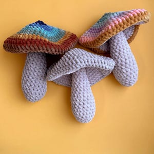 Pode incluir: Tr&ecirc;s brinquedos de cogumelo de croch&ecirc;. Um cogumelo &eacute; cinza, e os outros dois t&ecirc;m topos coloridos com uma base marrom.
