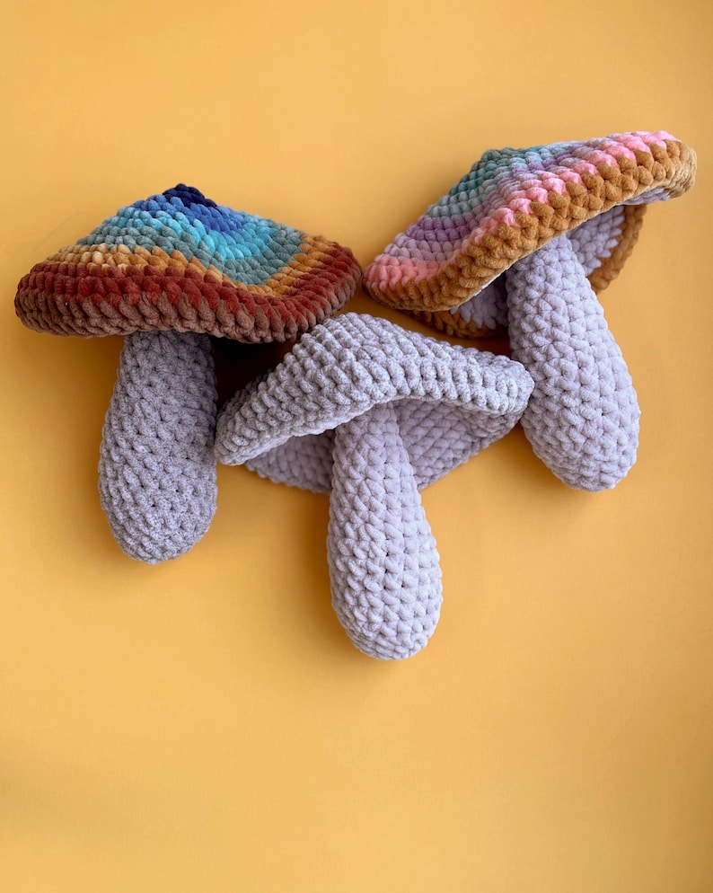 Pode incluir: Tr&ecirc;s brinquedos de cogumelo de croch&ecirc;. Um cogumelo &eacute; cinza, e os outros dois t&ecirc;m topos coloridos com uma base marrom.