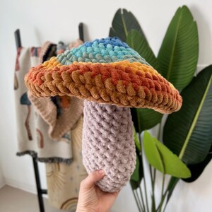 Pode incluir: Um cogumelo de croch&ecirc; arco-&iacute;ris com um chap&eacute;u marrom e um caule roxo. O cogumelo &eacute; feito com fio macio e &eacute; uma decora&ccedil;&atilde;o divertida e caprichosa para o quarto de uma crian&ccedil;a.