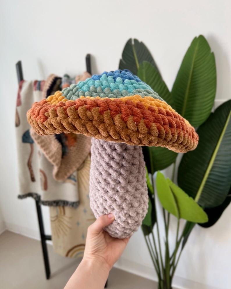 Pode incluir: Um cogumelo de croch&ecirc; arco-&iacute;ris com um chap&eacute;u marrom e um caule roxo. O cogumelo &eacute; feito com fio macio e &eacute; uma decora&ccedil;&atilde;o divertida e caprichosa para o quarto de uma crian&ccedil;a.
