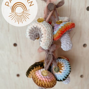 May include: A collection of handmade crochet mushroom ornaments in various colors, including pink, blue, and brown. Each mushroom has a unique design and is adorned with a small gold charm. The word "PATTERN" is displayed in a circular logo.