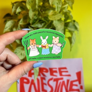 May include: A green sticker with the text "those who make peaceful revolution impossible will make violent revolution inevitable." The sticker features three cartoon animal characters holding a pitchfork, a bow tie, and a knife. Below the sticker is a sign that says "FREE PALESTINE!"