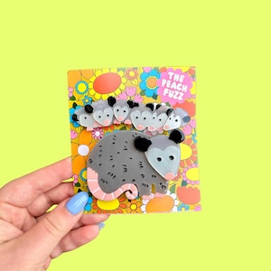 May include: A possum brooch with a gray body, pink tail, and black ears, accompanied by five smaller possums. The brooch is on a floral patterned card with the text "THE PEACH FUZZ" in a pink and orange bubble font.