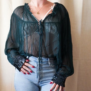 Op de afbeelding: Een doorschijnend groen blouse met een gerimpelde halslijn en lange mouwen. De mouwen hebben een zwarte paillettenbies. De blouse wordt gedragen over een zwarte kanten bh en gecombineerd met een blauwe jeans.