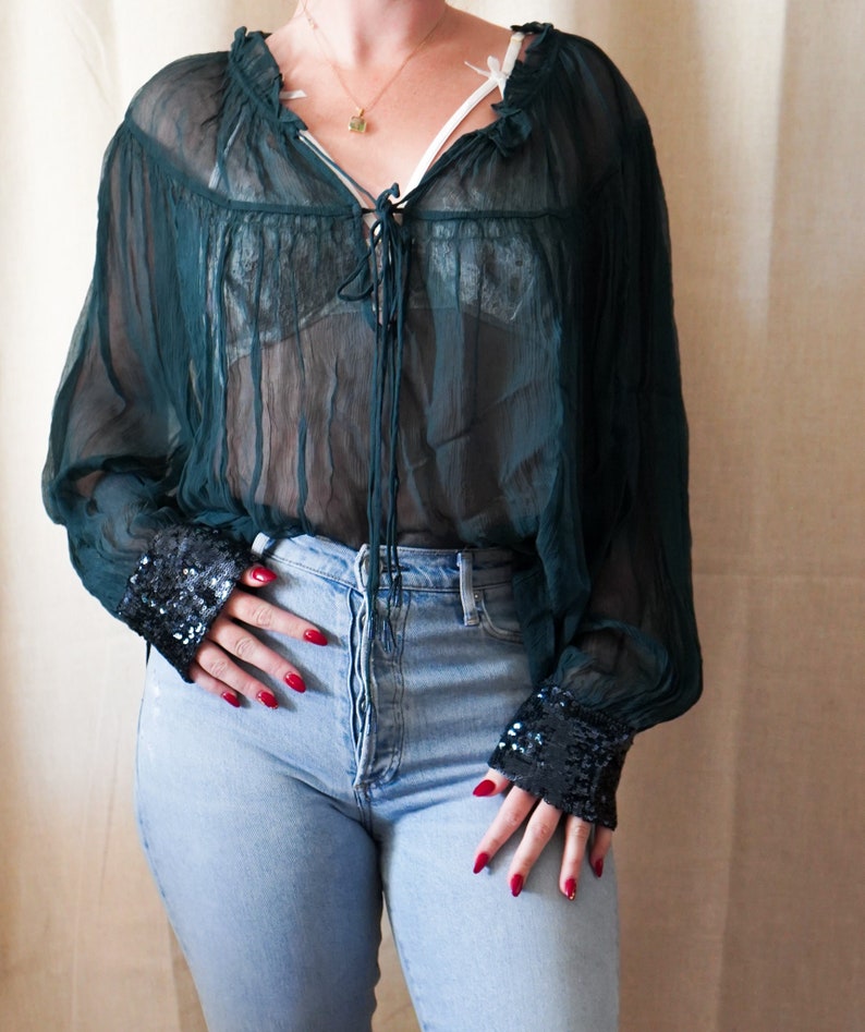 Op de afbeelding: Een doorschijnend groen blouse met een gerimpelde halslijn en lange mouwen. De mouwen hebben een zwarte paillettenbies. De blouse wordt gedragen over een zwarte kanten bh en gecombineerd met een blauwe jeans.
