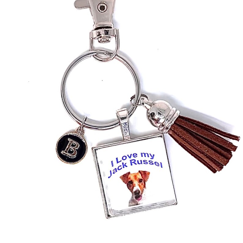 May include: A silver-toned metal keychain featuring a square pendant with a Jack Russell terrier illustration and the text I Love my Jack Russel. The keychain includes a small circular charm with the letter B and a brown faux leather tassel.