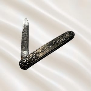 May include: A silver-toned metal nail file with a folding handle. The handle features an embossed, intricate scrollwork pattern across its surface. The file blade is textured and pointed, extending from the decorative metal casing at an angle.