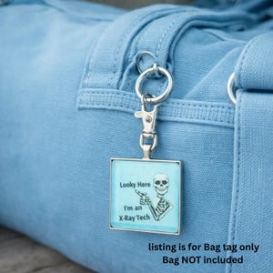 May include: A square bag tag with a silver-colored metal frame and a skeleton graphic on a light blue background. The tag reads "Looky Here, I'm an X-Ray Tech." The tag is attached to a light blue bag with a silver-colored clasp.