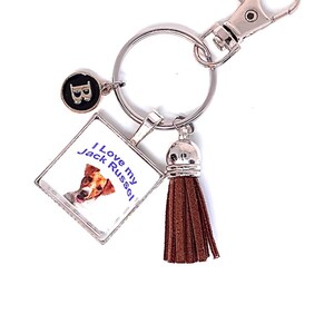 May include: A silver-toned metal keychain featuring a square charm with a Jack Russell terrier illustration and the text 'I Love my Jack Russel', a brown faux leather tassel, and a small circular charm with the letter 'B' in a black enamel inlay.