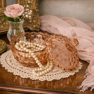 May include: A peach-colored, faceted glass jewelry box with a matching lid, filled with a strand of pearls. The box sits on a lace doily. A small vase with a pink rose and baby's breath is in the background.