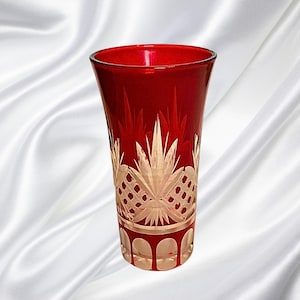 May include: A red glass vase with a detailed, etched design. The vase features a clear, decorative pattern near the base and a repeating design in the middle. The vase is made of red glass and has a smooth, polished finish.