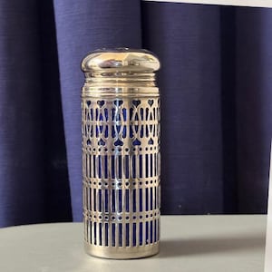 May include: A cylindrical glass shaker with a deep blue color, encased in a polished silver-tone metal sleeve featuring a decorative cut-out pattern of vertical bars and heart-shaped motifs, topped with a matching silver-tone metal lid.
