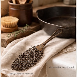 May include: A metal chainmail scrubber with a handle, resting on a beige cloth. The scrubber is designed for cleaning cookware. The text "listing is for Scrubber only" is visible at the bottom of the image.