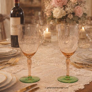 May include: Two elegant wine glasses with textured bowls and pink stems, resting on green bases. The glasses are set on a table with fine china, silverware, and a floral centerpiece, suggesting a formal dining setting.