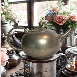 May include: A vintage silver teapot with a rounded body, curved spout, and a loop handle. The teapot sits atop a silver container, with other silver teapots and floral arrangements in the background. The scene evokes a sense of classic elegance and tea time.