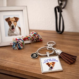 May include: A silver keychain featuring a square pendant with a Jack Russell terrier image and the text I Love my Jack Russel, a small round charm with the letter B, and a brown faux leather tassel attached to a metal split ring.