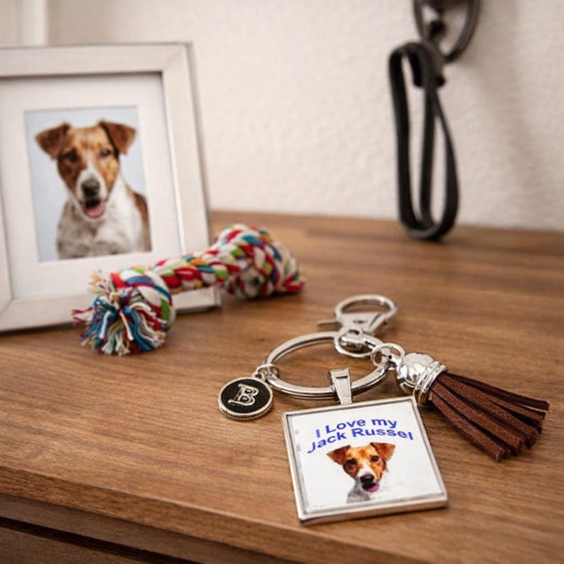 May include: A silver keychain featuring a square pendant with a Jack Russell terrier image and the text I Love my Jack Russel, a small round charm with the letter B, and a brown faux leather tassel attached to a metal split ring.