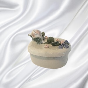 May include: A small, oval-shaped ceramic trinket box with a lid. The lid is decorated with a pink rose, green leaves, and small pink and blue floral accents. The box is a light beige color.
