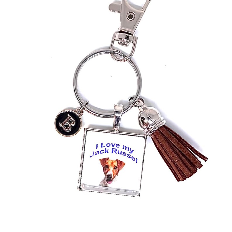 May include: A silver-toned metal keychain featuring a square pendant with a Jack Russell terrier illustration and the text I Love my Jack Russel, a small circular charm with the letter B, and a brown faux leather tassel attached to a split ring.
