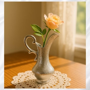 May include: A small, silver-colored pitcher with a handle, holding a peach-colored rose. The pitcher sits on a wooden table with a cream-colored lace doily. The background includes a window with sheer curtains.
