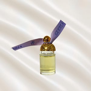 May include: A small clear glass perfume bottle filled with pale yellow liquid. The bottle features a gold-colored cap with a textured honeycomb pattern and a light purple ribbon tied around the neck, printed with the text GUERLAIN.