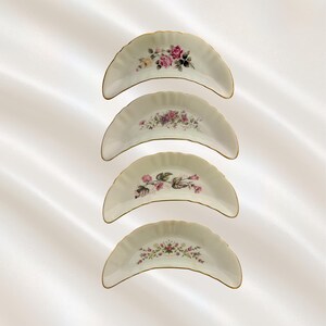 May include: A set of four crescent-shaped porcelain bone dishes with scalloped edges and gold trim. Each dish features a unique floral pattern in shades of pink, green, and purple, arranged centrally on the white surface of the curved plates.