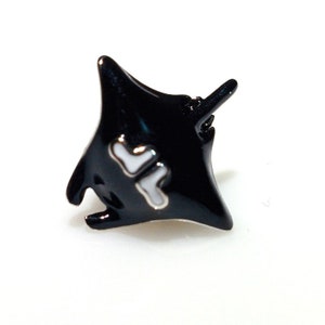 Pode incluir: Um broche de arraia manta preto com detalhes brancos. A arraia manta está em posição de voo com as asas abertas.