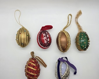May include: A collection of six decorative Easter egg ornaments. The eggs feature various colors, textures, and embellishments, including gold, red, green, and silver. Some are adorned with beads, sequins, and ribbons, while others have metallic finishes. Each egg has a loop for hanging.