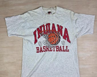 Könnte beinhalten: Hellgraues T-Shirt mit dem roten Schriftzug "INDIANA BASKETBALL" und einem Basketball, der durch ein Netz geht. Das Shirt hat kurze Ärmel und einen Rundhalsausschnitt.