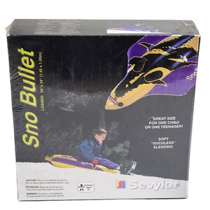 Puede incluir: Una caja de trineo Sno Bullet, 142cm x 100cm, con un niño montado. La caja presenta un gráfico de trineo morado y amarillo y el texto "Great size for one child or one teenager!" y "Soft 'ouchless' sledding."