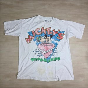 May include: White t-shirt with a graphic of Mickey and Minnie Mouse in a pink boat, with the text "Mickey's Wavemaker". The design includes a blue triangle and green text. The shirt is vintage and shows signs of wear.